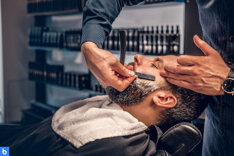 This is the cover for our Best Straight Razor article. It features a man sitting in a barber shop being shaved with a straight razor. The background looks like a barber shop, but out of focus in the image.