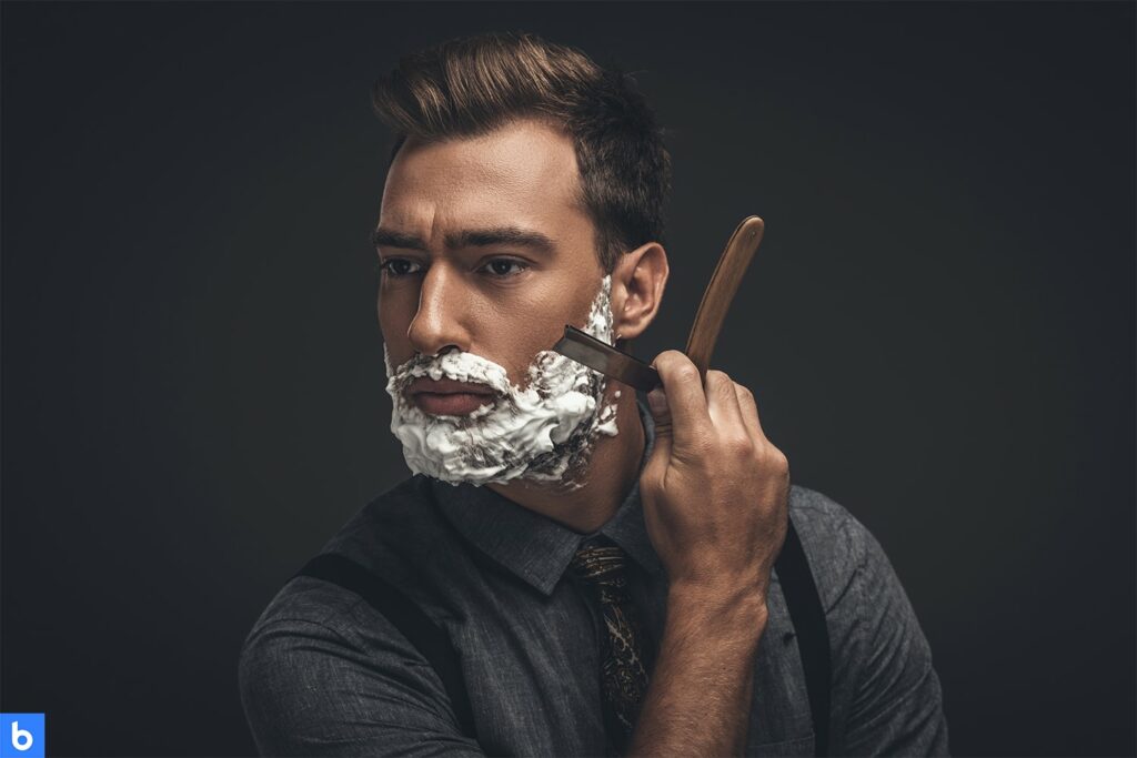 This is a cover photo for the Best Straight Razor 2024 Buying Guide. It is a photo of a man using shaving cream and a straight razor to shave. He is dressed in a slate grey shirt with a tie and suspenders, with a dark black background behind him.