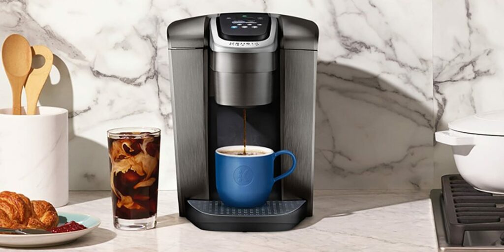 This is a photo of a brushed gunmetal silver Keurig single serve coffee maker with a blue cup placed on a marble counter top. Cup of espresso coffee placed on the counter. Plate with pastry and a jar of utensils in the background. 
