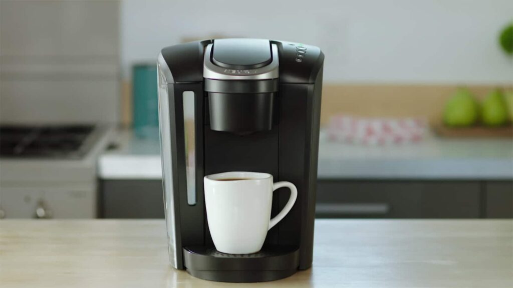 This is the feature photo for our Best Single Serve Coffee Maker 2024 article. It features a black Keurig coffee maker with a white cup sitting on a kitchen counter with blurry background.