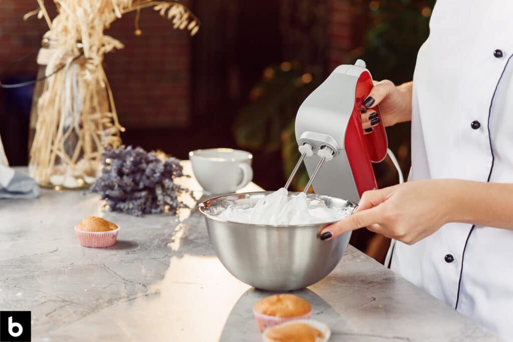 This is a photo for the Best Hand Mixer 2024 article. Woman mixing whipped cream in a mixing bowl with a hand mixer. There are several muffins sitting on the counter in the background.