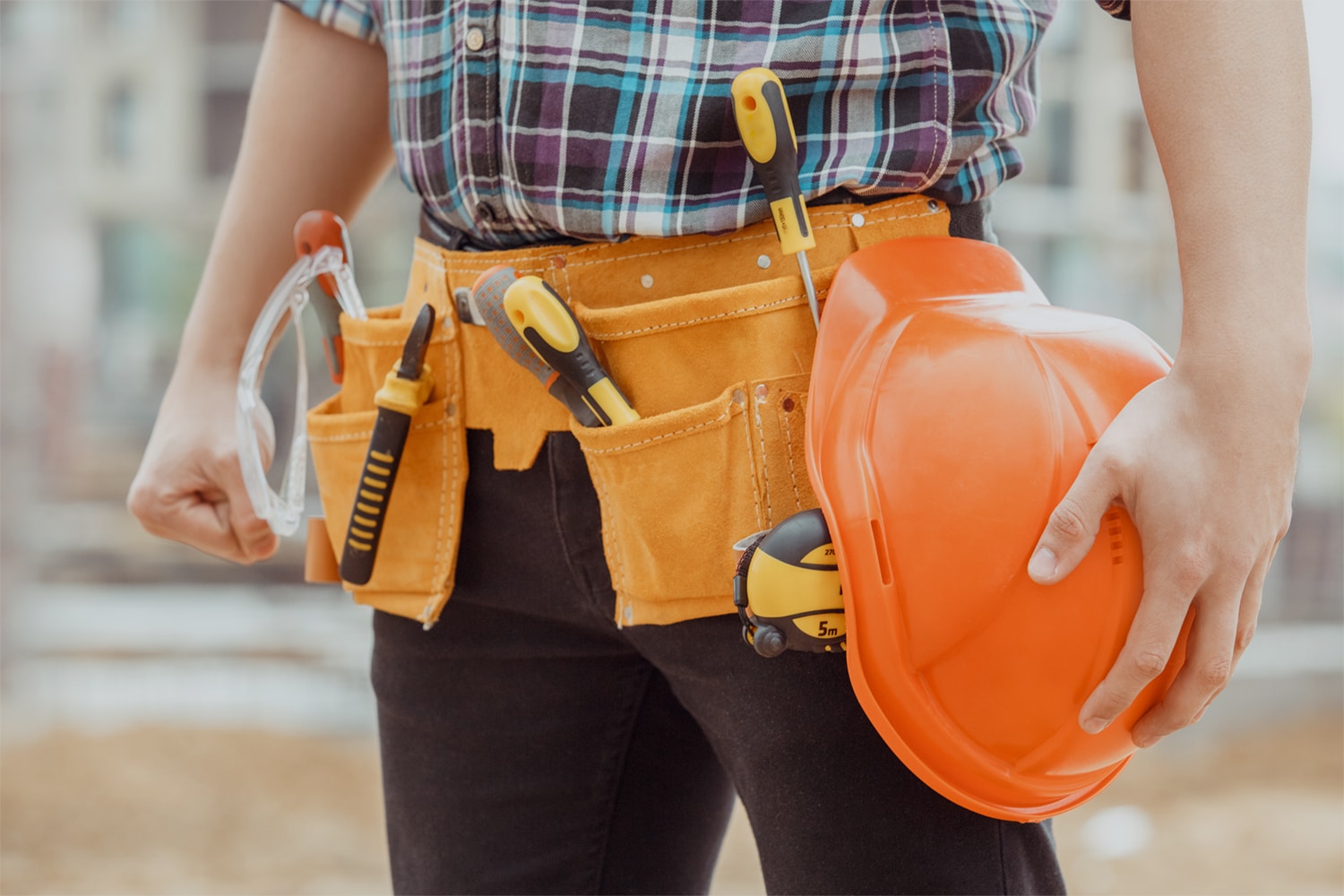 This is the cover photo for our Best Tool Belt article. It shows a construction worker wearing black pants and a plaid shirt wearing a tool belt with assorted tools inside. There is a blurred out background of the construction site.