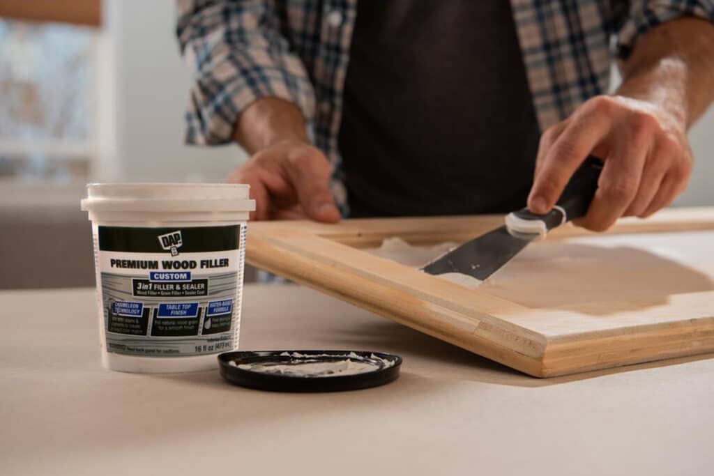 This is a cover photo for our Wood Filler Buying Guide 2024. It shows a person applying wood filler to a cabinet door with a spatula. They are wearing a plaid shirt and there is a bucket of wood filler placed on the table. There is a blurry garage background behind the person.