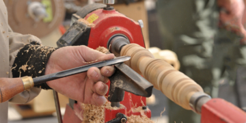 This is the cover photo for our Best Wood Lathe article. It shows a person using a wood lathe to shave and shape a wood dowel. There are wood shavings flying all over, and a blurred background with various tools in the peripheral.