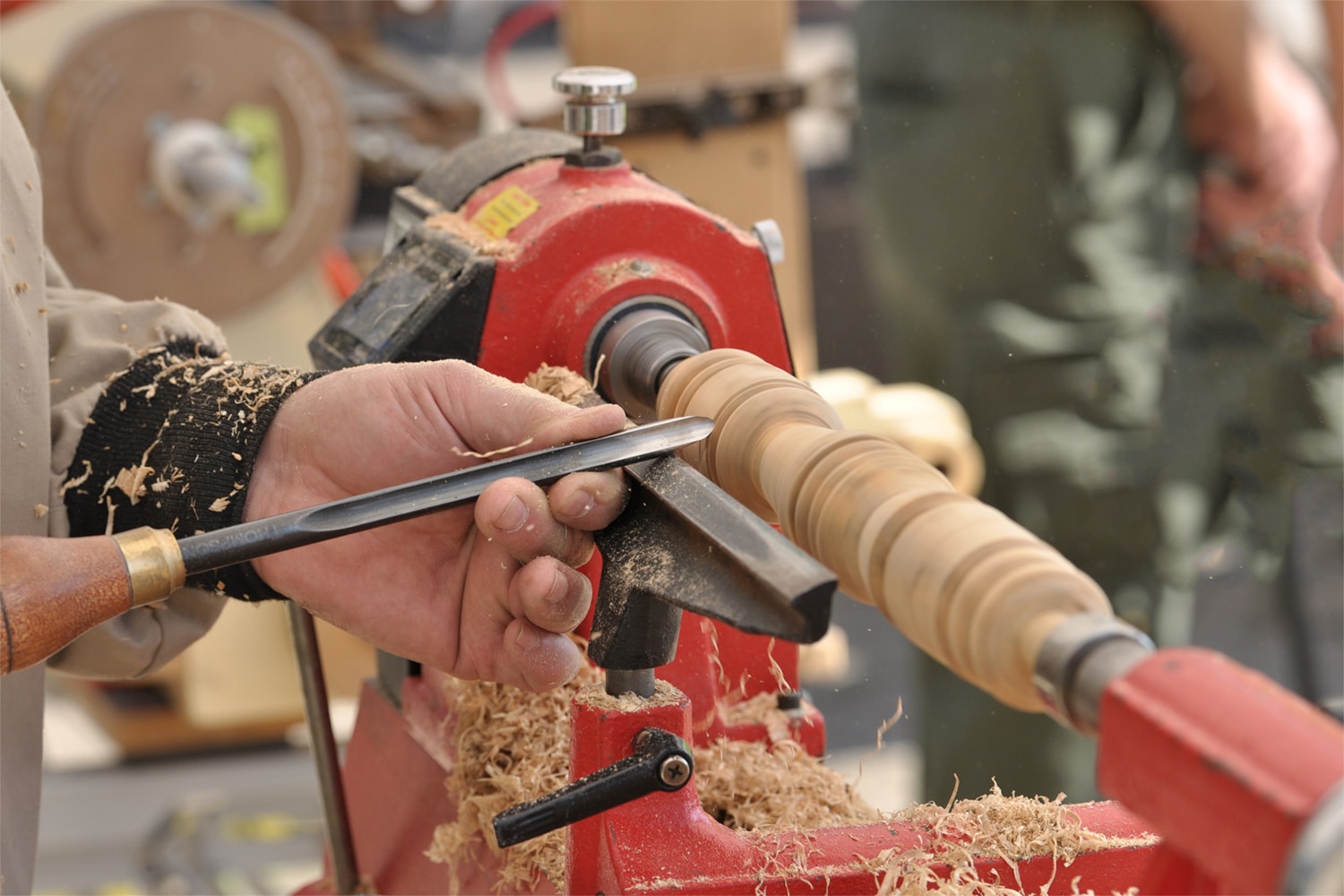 This is the cover photo for our Best Wood Lathe article. It shows a person using a wood lathe to shave and shape a wood dowel. There are wood shavings flying all over, and a blurred background with various tools in the peripheral.