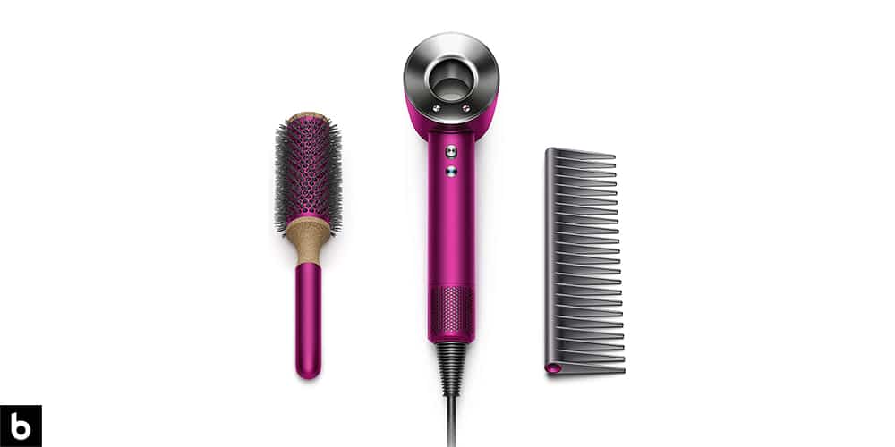 This is a product image in our Best Hair Dryers for Curly Hair in 2024 article. It is a photo of a Dyson Supersonic Hair Dryer overlaid on a minimalistic white background with a Burbro logo. There is a brush and comb in the photo as well.