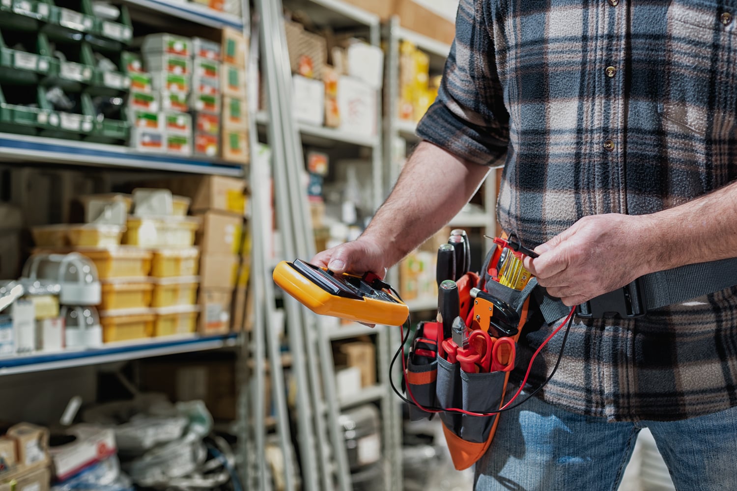 This is the cover photo for our Best Electrician Tool Belt article. It shows an electrician in a store isle, holding a multimeter, wearing a tool belt with tools inside. The background has several shelves of supplies.
