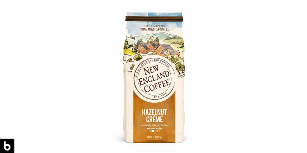 This is a product image for our Best Flavored Coffee 2024 article. It features a blue and gold colored bag of New England Hazelnut Crème coffee beans. There is a watercolor painting of a countryside on the side of the bag.