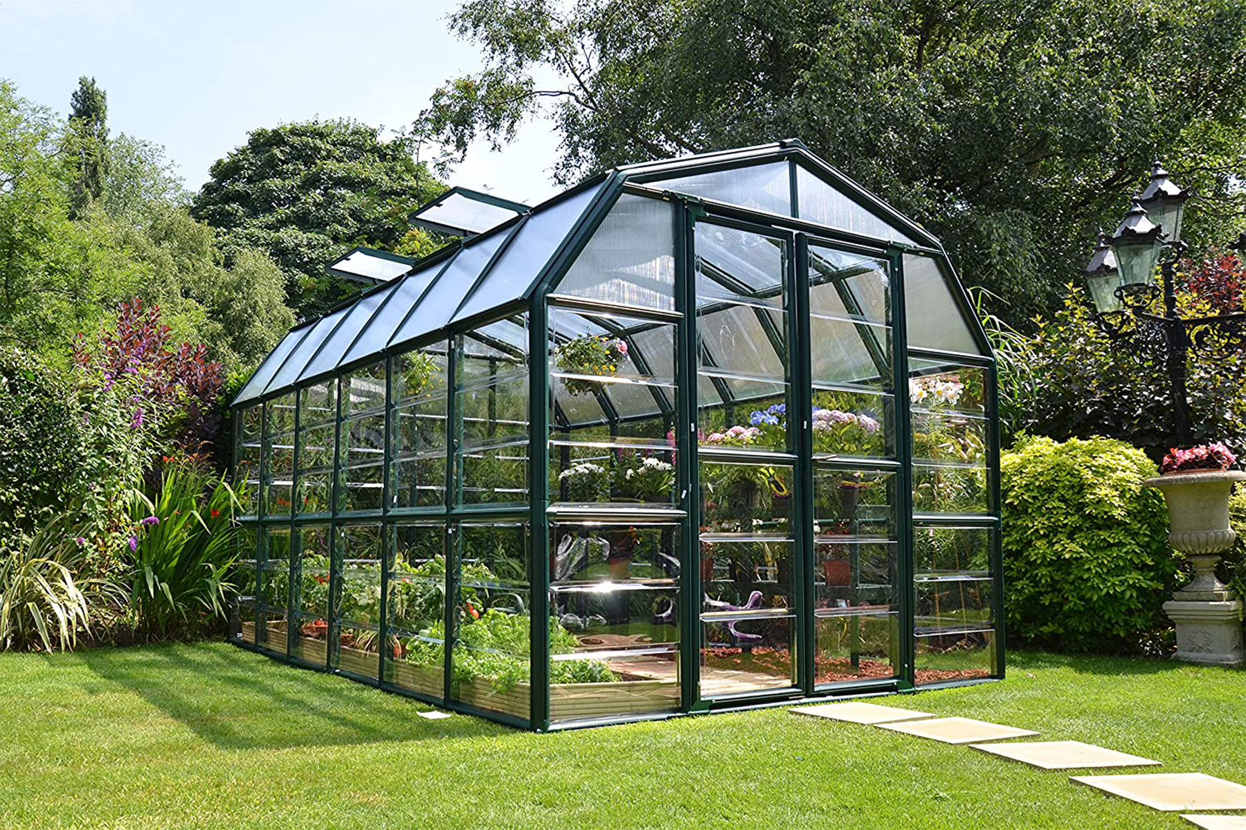 This is the cover photo for our Best Greenhouse Kits article. It features a greenhouse with plants inside, situated in a garden with green grass, trees, bushes, and flowers.