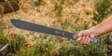 This is the cover photo for our Best Machetes article. It features a man's arm holding a metal machete with grass and trees in the background.