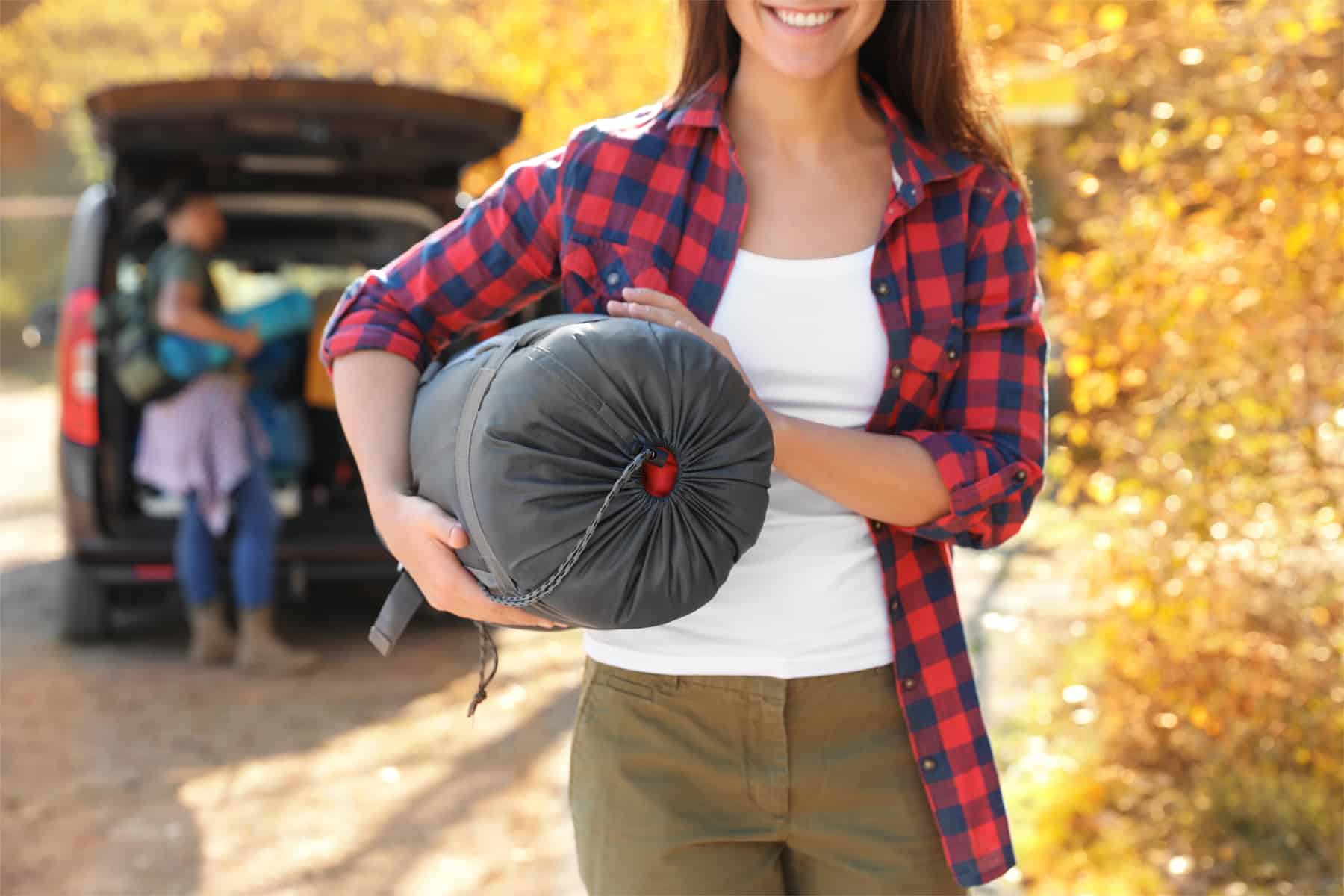 This is the cover photo for our Best Sleeping Bag for Camping article. It features a woman holdings a sleeping bag with a man loading a vehicle in the background.