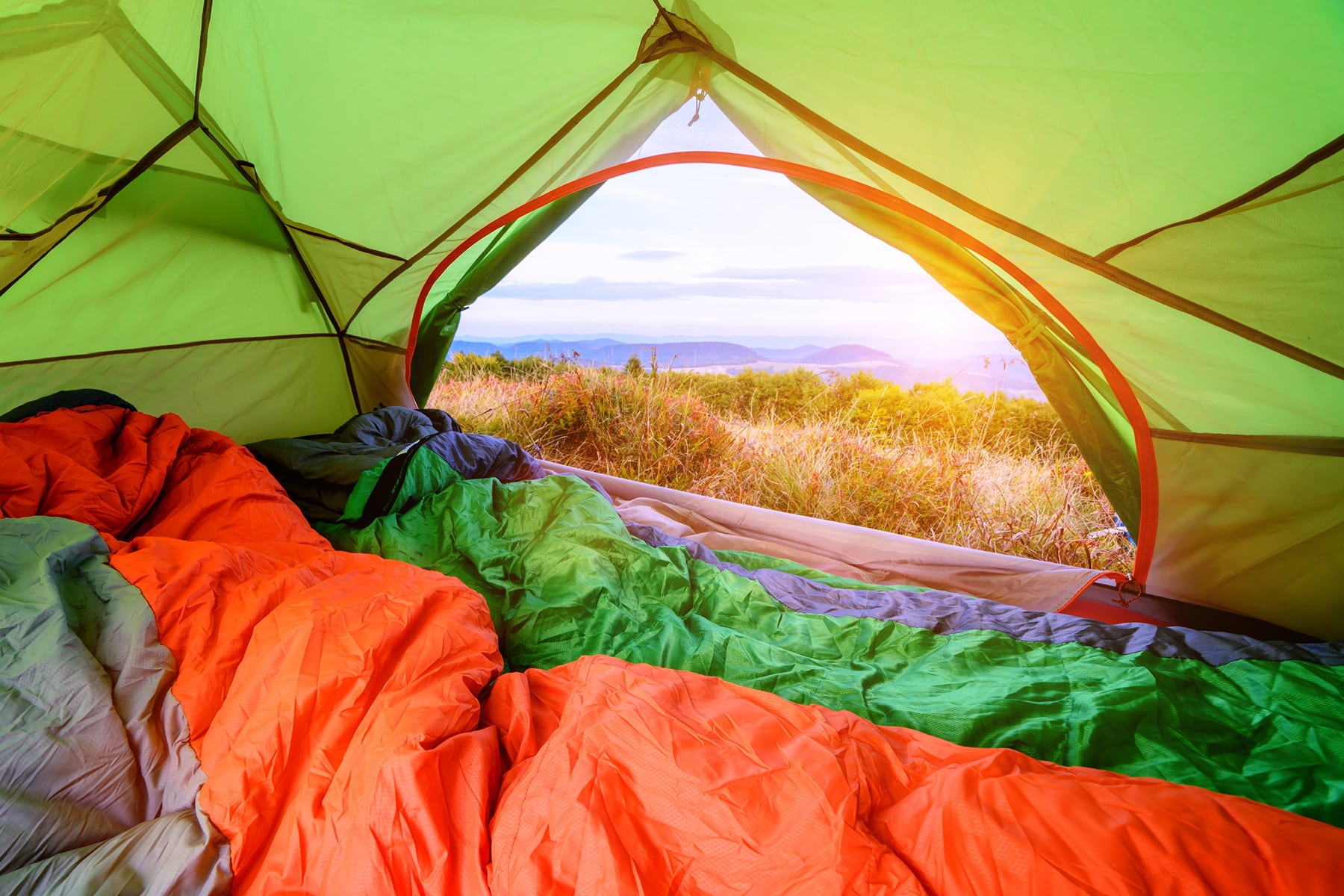 This is the cover photo for our Best Sleeping Bags articles. It features orange and green sleeping bags inside a green tent, with the front flap open to a scenic view outside.